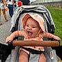 baby, stroller, hat, smiling, happy, outdoor, people, sidewalk, grass, clothing, sunhat, child, infant, walking, family, daylight, road, casual, person, fun