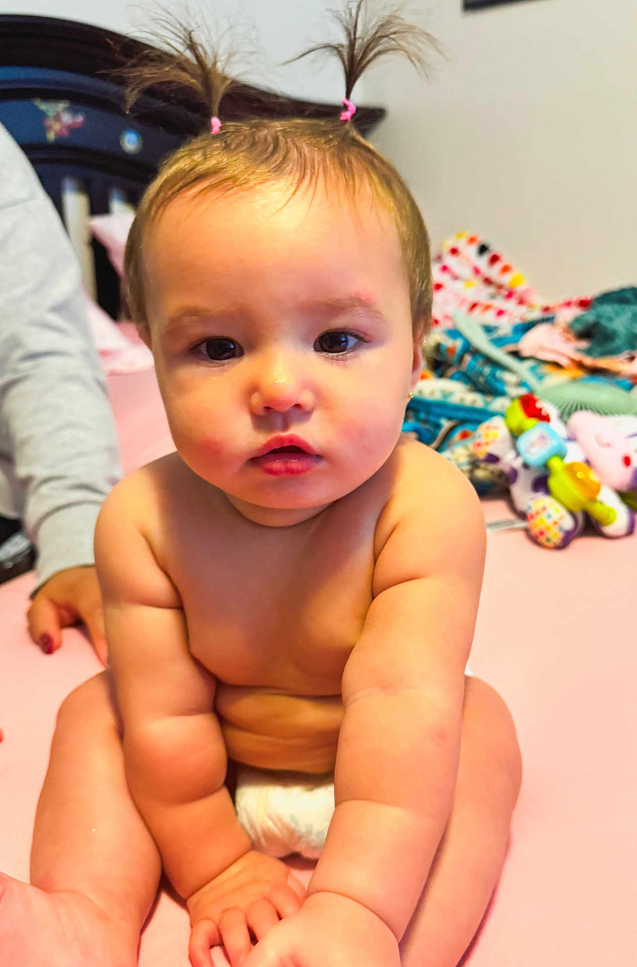 Rilynn is registered to the contest to win money with this photo: baby, child, pigtails, pink_bed, diaper, toys, blanket, hand, curious, indoor, cute, chubby, person, skin, face, sitting, infant, young_child, bedroom, soft_lighting