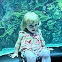 child, blonde_hair, floral_outfit, sitting, bench, aquarium, coral, marine_life, underwater, curious, indoor, portrait, young, pink, green, sea_creatures, water, face, person, light