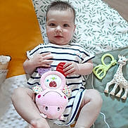Rosie participe au concours pour gagner de l'argent avec cette photo : baby, child, toy, plush_toy, rattle, giraffe_toy, striped_dress, play_mat, smiling, cute, infant, lying_down, hands, feet, indoor, floor, soft_toy, colorful, patterned, happy
