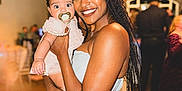 Skyler is registered to the contest to win money with this photo: woman, baby, pacifier, braids, smile, indoor, event, people, dress, holding, happy, face, person, blurred_background, party, skin, hair, child, celebration, warm_lighting