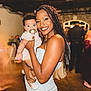woman, baby, pacifier, braids, smile, indoor, event, people, dress, holding, happy, face, person, blurred_background, party, skin, hair, child, celebration, warm_lighting