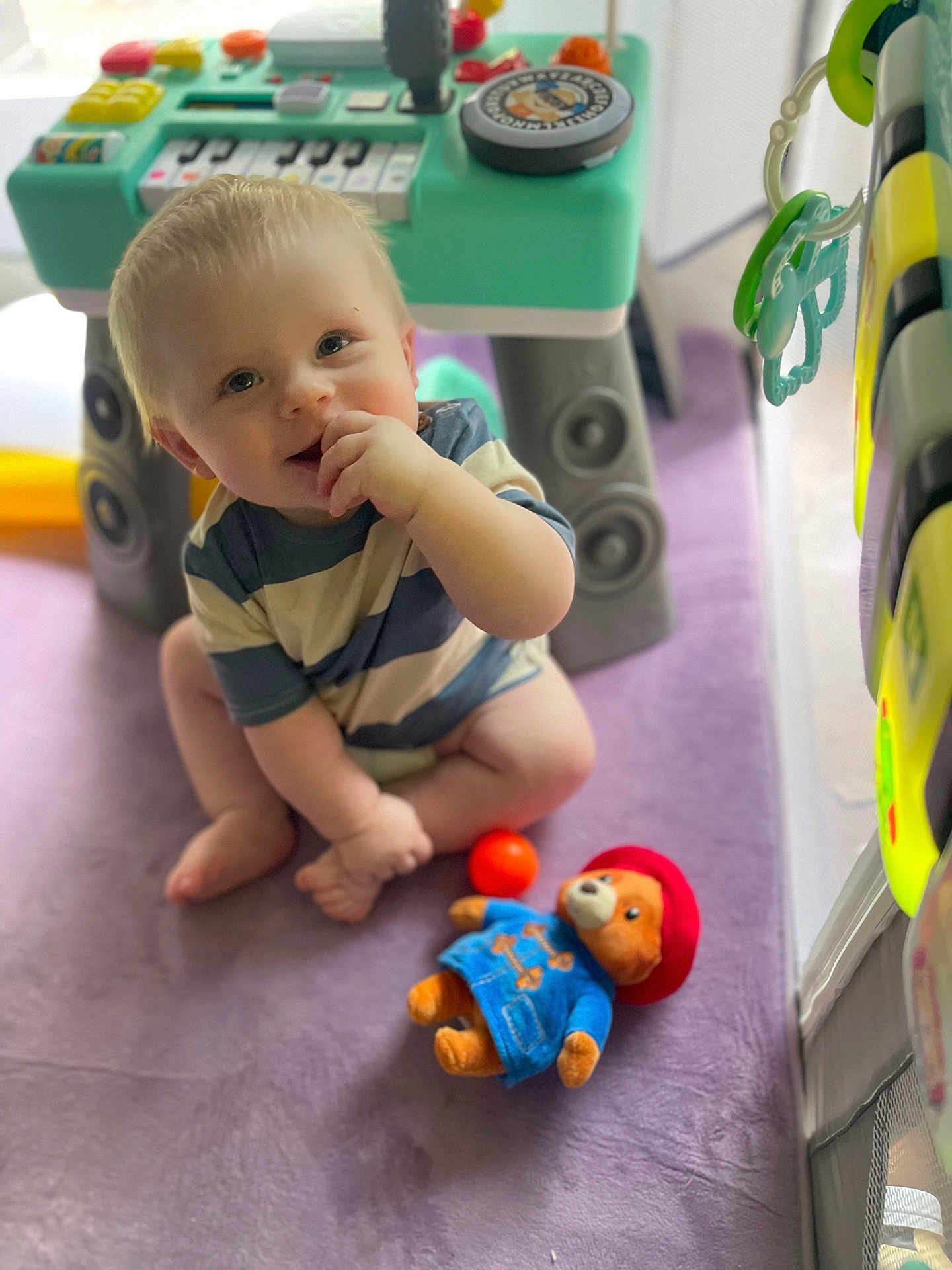 Rayland joined the competition — help win amazing prizes! baby, child, toy, plush_bear, keyboard_toy, purple_blanket, indoor, play_area, cute, smiling, sitting, striped_shirt, soft_light, floor, hand, foot, blond_hair, face, happy, playtime