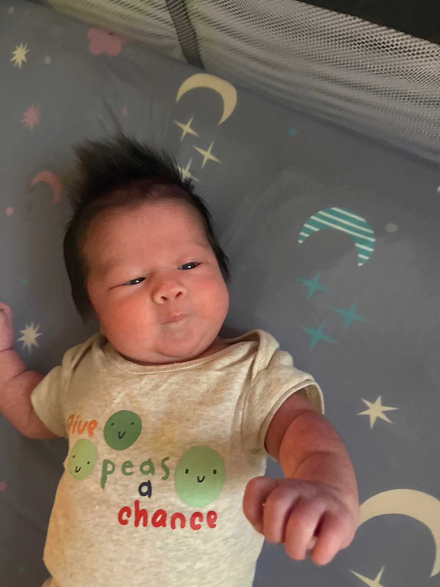 Ryley is registered to the contest to win money with this photo: baby, infant, child, cute, clothing, shirt, text, peas, fist, face, hair, lying_down, blanket, stars, moon, pattern, expression, indoors, newborn, person