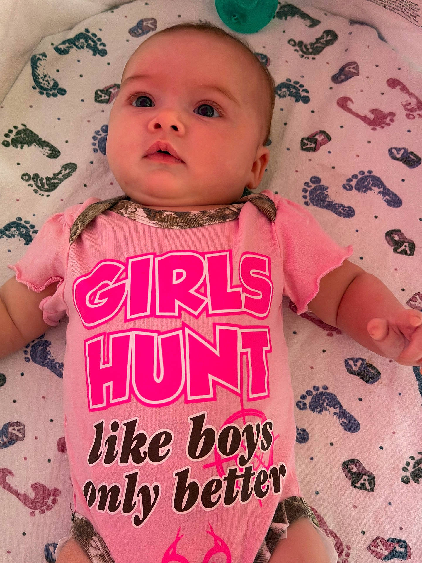 baby, baby_clothing, blanket, casual_photo, child, close_up, face, green_toy, hands, indoor, infant, lying_down, onesie, patterned_blanket, pink_outfit, playful, portrait, smiling, text_on_clothing, wide_eyed