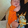 Leonidas joined the competition — help win amazing prizes! baby, newborn, sleeping, infant, orange_outfit, portrait, close_up, peaceful, resting, adult, parent, cuddling, cartoon_shirt, dog_character, indoors, blanket, hand, arm, soft_lighting, family