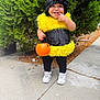 child, toddler, halloween, bee_costume, pumpkin_bucket, smile, laughing, outdoors, evergreen_tree, sidewalk, sneakers, white_shoes, black_clothing, yellow_fur, hat, trick_or_treat, cute, standing, portrait, candy_bucket
