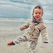 Yanael a rejoint le concours — aidez-le/la à gagner de superbes lots ! toddler, child, beach, sand, ocean, cloudy_sky, jacket, pants, shoes, playful, smiling, curly_hair, outdoor, pose, waves, water, fun, happy, nature, cozy