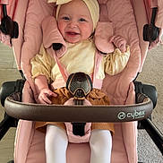 Fidjy participe au concours pour gagner de l'argent avec cette photo : baby, stroller, pink, flower_headband, boots, smiling, child, indoor, cozy, cute, tights, yellow, brown_skirt, seatbelt, wheels, carriage, happy, infant, person, portrait