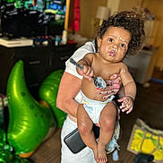Castiel is registered to the contest to win money with this photo: adult_hand, baby, curly_hair, diaper, dinosaur, expression, floor, food, footwear, furniture, indoor, inflatable, living_room, messy, nail_polish, person, skin, spoon, television, toy