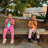 Castiel is registered to the contest to win money with this photo: toddler, child, bench, outdoor, sitting, pink_clothing, orange_clothing, shoes, drink, cup, trees, building, sunlight, playful, face, person, nature, leaves, grass, side_by_side
