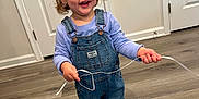 Valentina joined the competition — help win amazing prizes! toddler, child, smiling, indoor, denim_overalls, barefoot, curly_hair, pony_tail, wooden_floor, white_door, holding_string, long_sleeve_shirt, happy, person, flooring, cute, clothing, playful, young_child, standing