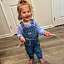 toddler, child, smiling, indoor, denim_overalls, barefoot, curly_hair, pony_tail, wooden_floor, white_door, holding_string, long_sleeve_shirt, happy, person, flooring, cute, clothing, playful, young_child, standing