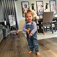 Valentina is registered to the contest to win money with this photo: toddler, child, barefoot, denim_overalls, curly_hair, balloon, wooden_floor, living_room, dining_table, chairs, high_chair, curtains, plush_toy, framed_photos, portrait, indoor, person, home, floor, string