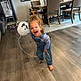 Valentina is registered to the contest to win money with this photo: toddler, child, balloon, indoor, barefoot, denim_overalls, curly_hair, happy, smiling, wooden_floor, dining_room, chair, table, rug, home_interior, playful, casual_clothing, person, young_child, joyful