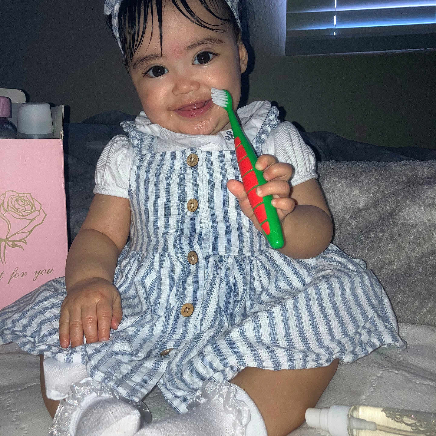Daynelis is registered to the contest to win money with this photo: baby, child, smiling, toothbrush, striped_dress, headband, sitting, socks, indoor, bed, blanket, window_blinds, cute, happy, person, infant, hand, footwear, portrait, playful
