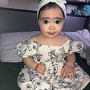 Daynelis is registered to the contest to win money with this photo: baby, child, face, headband, dress, flower_pattern, sitting, bed, pillow, plush_toys, stuffed_animals, indoor, cute, portrait, hands, feet, expression, young_child, clothing, person