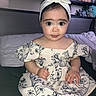 baby, child, face, headband, dress, flower_pattern, sitting, bed, pillow, plush_toys, stuffed_animals, indoor, cute, portrait, hands, feet, expression, young_child, clothing, person