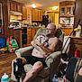 armchair, baby, baby_bottle, bottle, cabinet, clothing, family, furniture, glasses, holding, indoor, kitchen, living_room, man, person, prosthetic_leg, refrigerator, table, tattoos, wooden_floor