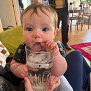 Aria is registered to the contest to win money with this photo: baby, infant, child, glass, ice, drinking, tongue, hands, face, eyes, person, adult, living_room, sofa, chair, carpet, indoor, cozy, curious, holding