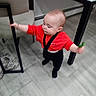 Dolly is registered to the contest to win money with this photo: toddler, child, red_shirt, black_suspender, standing, indoor, chair, table, floor, holding, leafy_plant, baby, person, clothing, furniture, home, cute, small_child, young, blurred