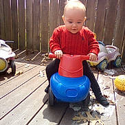 Dolly is registered to the contest to win money with this photo: toddler, child, toy, ride_on_toy, red_sweater, outdoor, wooden_deck, leaves, play, baby, plastic_toy, concentration, sunlight, casual_clothing, footwear, fence, daytime, person, cute, fun