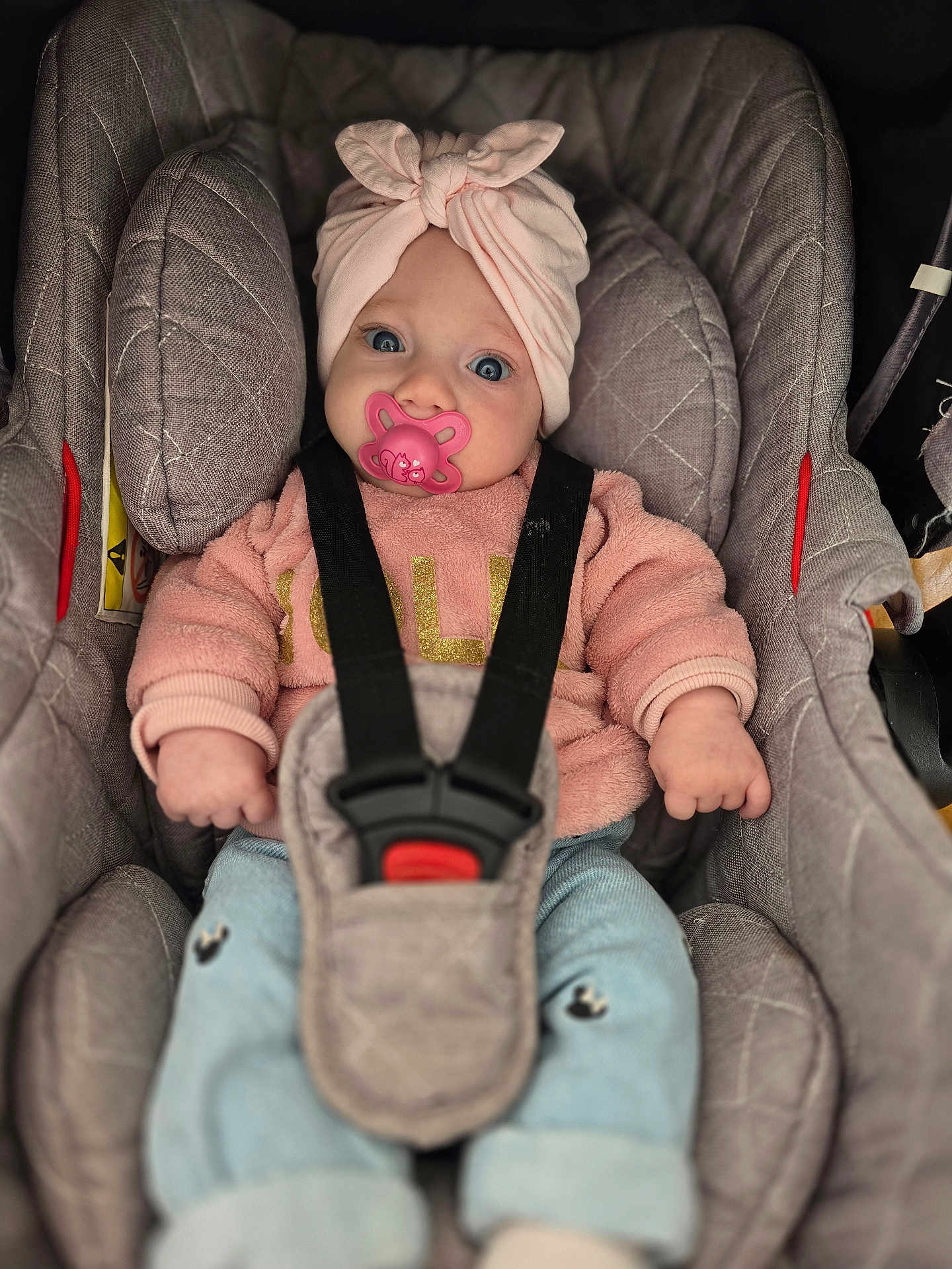 Kiara a rejoint le concours — aidez-le/la à gagner de superbes lots ! baby, infant, car_seat, pacifier, headwrap, pink_clothing, sweater, blue_eyes, child, straps, safety_harness, cute, portrait, indoor, seat, clothing, person, baby_girl, cozy, closeup