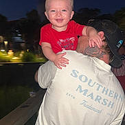Rylan is registered to the contest to win money with this photo: baby, child, adult, smiling, red_shirt, white_shirt, cap, tattoo, night, outdoor, portrait, person, happiness, holding, face, hand, joy, blurred_background, casual_clothing, people