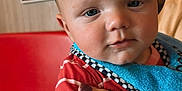 Thomas is registered to the contest to win money with this photo: baby, child, burger_king, crown, red_shirt, striped_clothing, hand, adult_hand, fast_food, restaurant, booth, table, blue_bib, face, skin, portrait, indoor, seated, cute, expression