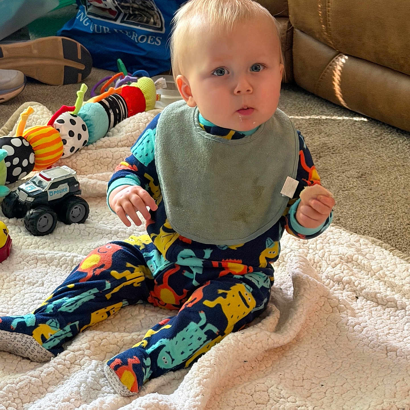 Nolan joined the competition — help win amazing prizes! baby, child, infant, bib, pajamas, dinosaur_print, toy, toy_car, blanket, carpet, couch, indoor, floor, face, blue_eyes, sitting, playful, colorful, cute, home