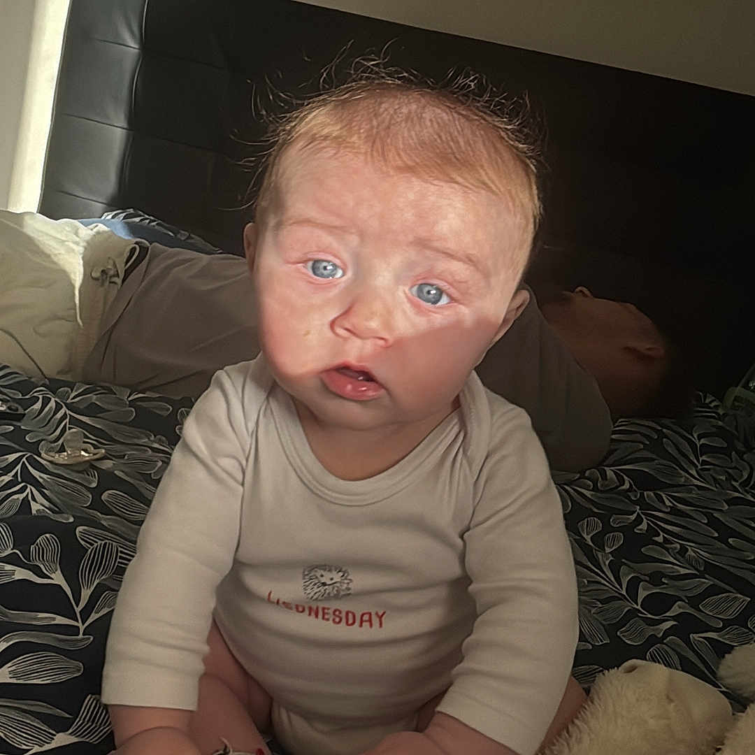 Elio Briche participe au concours pour gagner de l'argent avec cette photo : adult_in_background, baby, bed, bedding, blanket, blue_eyes, child, hands, headboard, indoor, infant, onesie, pacifier, pattern, plush_toy, portrait, shadow, sitting, socks, sunlight