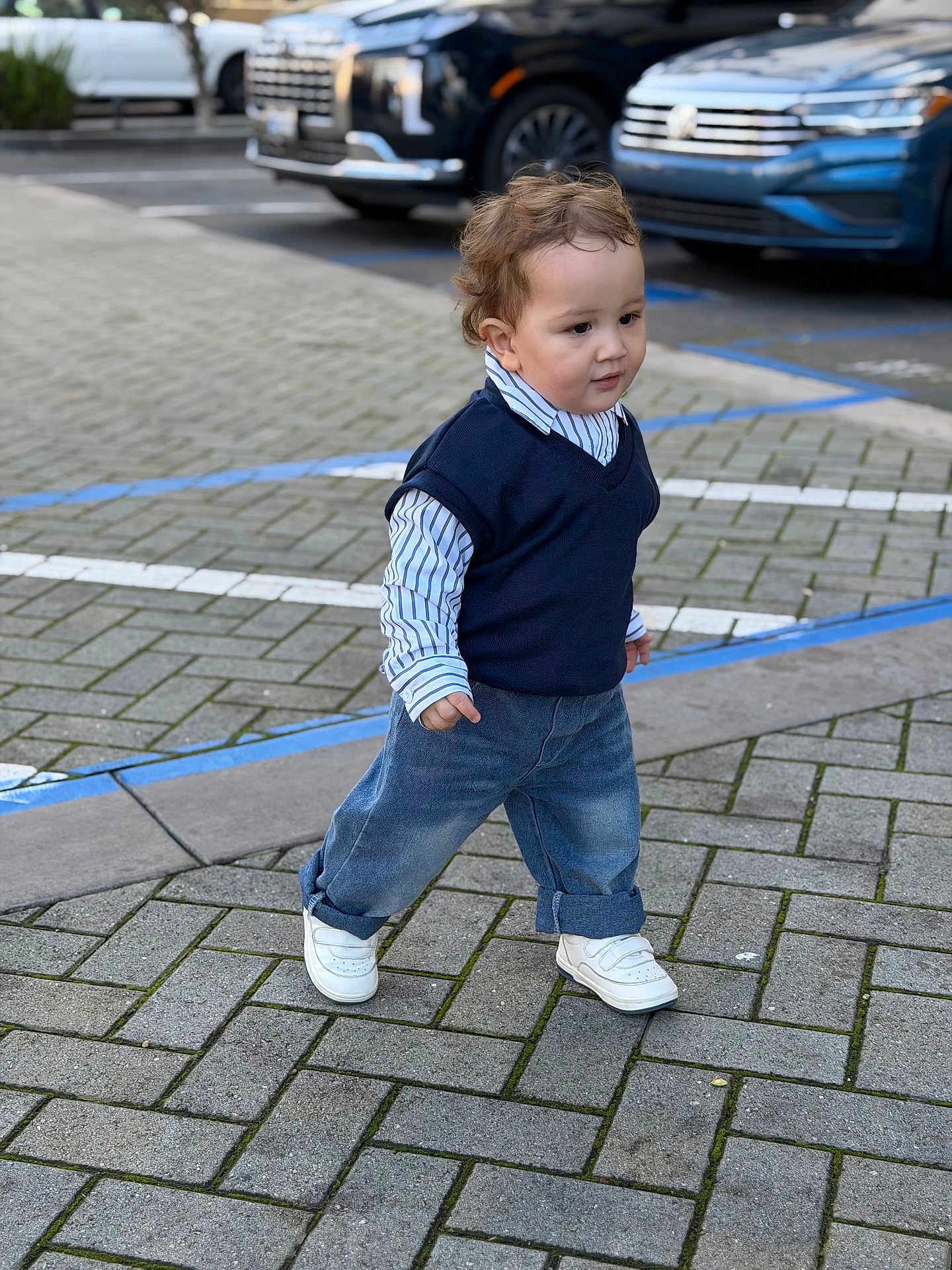 Yanis is registered to the contest to win money with this photo: toddler, child, walking, street, pavement, jeans, shirt, vest, sneakers, cars, parking_lot, outdoor, curb, sidewalk, person, fashion, casual, young_child, daytime, urban