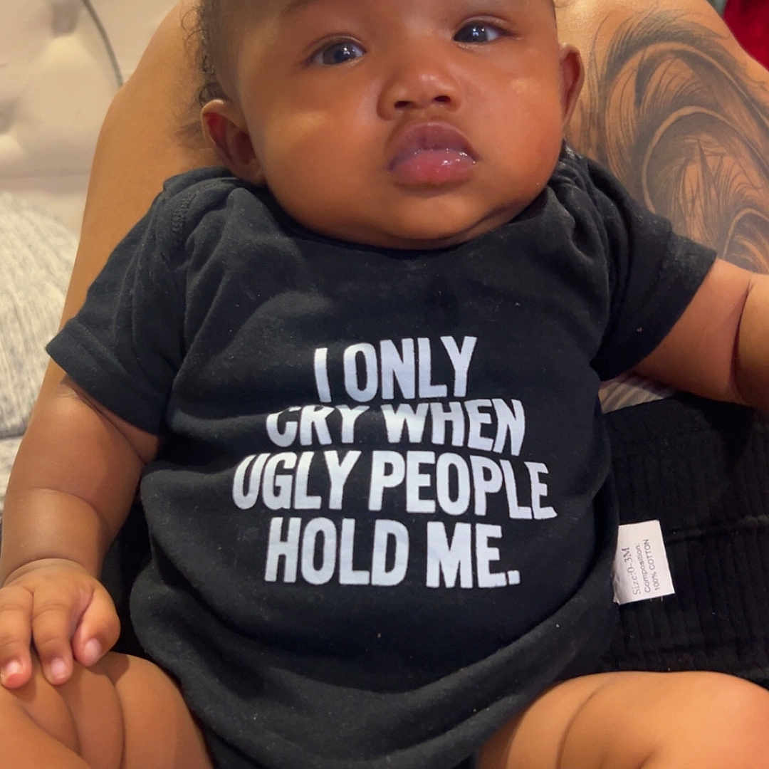 Kamali joined the competition — help win amazing prizes! baby, child, onesie, sitting, tattoo, indoor, furniture, person, clothing, expression, cute, skin, hand, footwear, sock, humor, text_on_clothing, black_clothing, infant, portrait