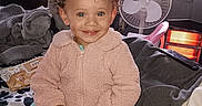 Neveah is registered to the contest to win money with this photo: air_conditioner, bed, blanket, child, clothing, curly_hair, curtain, face, fan, furniture, happy, heater, indoor, jacket, pants, person, room, smiling, toddler, window