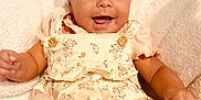 Malia is registered to the contest to win money with this photo: baby, child, smiling, infant, floral_dress, headwear, white_turban, blanket, happy, cute, portrait, skin, person, indoors, baby_girl, clothing, soft_texture, button, hands, lying_down