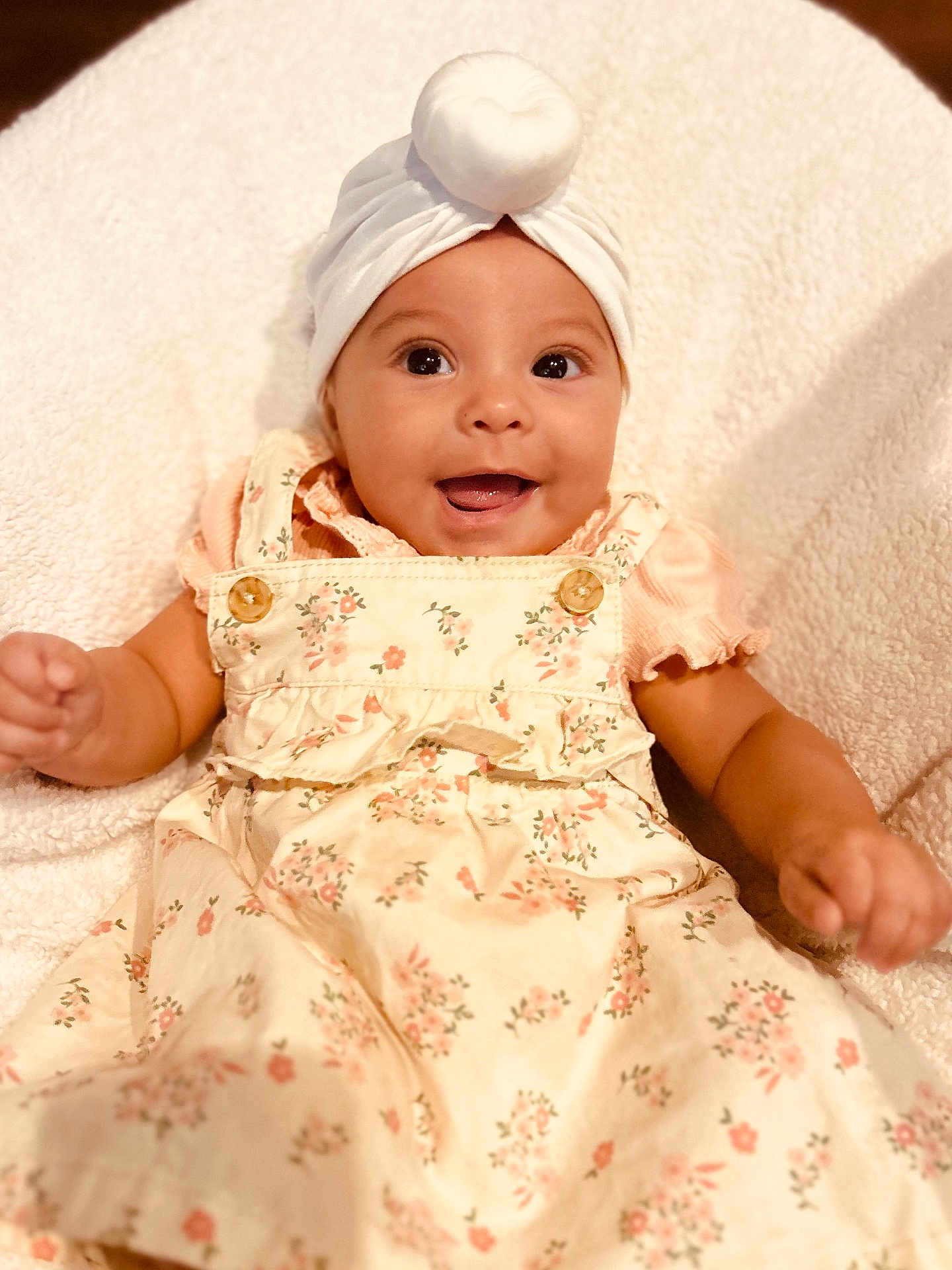 Malia is registered to the contest to win money with this photo: baby, child, smiling, infant, floral_dress, headwear, white_turban, blanket, happy, cute, portrait, skin, person, indoors, baby_girl, clothing, soft_texture, button, hands, lying_down