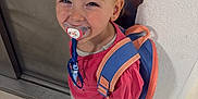 Maddy participe au concours pour gagner de l'argent avec cette photo : child, pacifier, backpack, smile, standing, floor, tile, wall, door, footwear, pink_shirt, blonde_hair, happy, indoor, young_child, casual_clothing, playful, person, cute, innocent