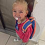 child, pacifier, backpack, smile, standing, floor, tile, wall, door, footwear, pink_shirt, blonde_hair, happy, indoor, young_child, casual_clothing, playful, person, cute, innocent