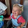 toddler, child, smiling, laughing, blonde_hair, pigtails, hair_bow, denim_overalls, red_shirt, high_chair, cup, indoor, window, curtains, wooden_door, glass_table, adult_partial, happy, person, seated