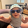 action_figure, baby, child, floor_tiles, furniture, hand, hat, indoor, joyful, living_room, person, playful, portrait, shorts, skin, smiling, sunglasses, tank_top, toddler, window