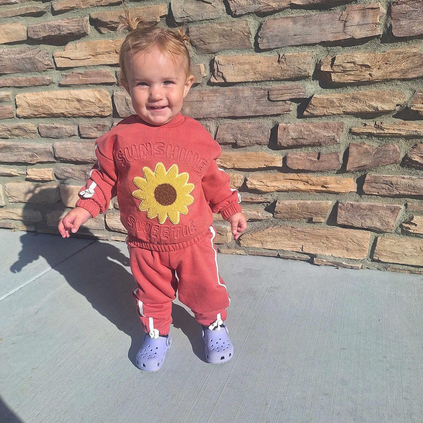 Harmony joined the competition — help win amazing prizes! toddler, child, smiling, standing, outdoor, sunlight, sunflower, sweatshirt, tracksuit, red_clothing, purple_shoes, clogs, brick_wall, stone_wall, patio, shadow, portrait, full_body, happy, fashion