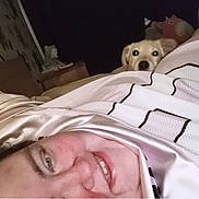 Floki is registered to the contest to win money with this photo: person, dog, bed, blanket, pillow, smile, face, indoor, nighttime, dark, curious, peeking, furniture, room, head, comfort, sleepwear, cute, pet, relaxation