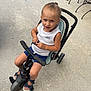 Tonio participe au concours pour gagner de l'argent avec cette photo : toddler, child, tricycle, outdoor, concrete, curious, blue_shorts, white_tank_top, sandals, small_pony_tail, toy, seat, handlebars, feet, expression, summer, playtime, person, casual, daylight