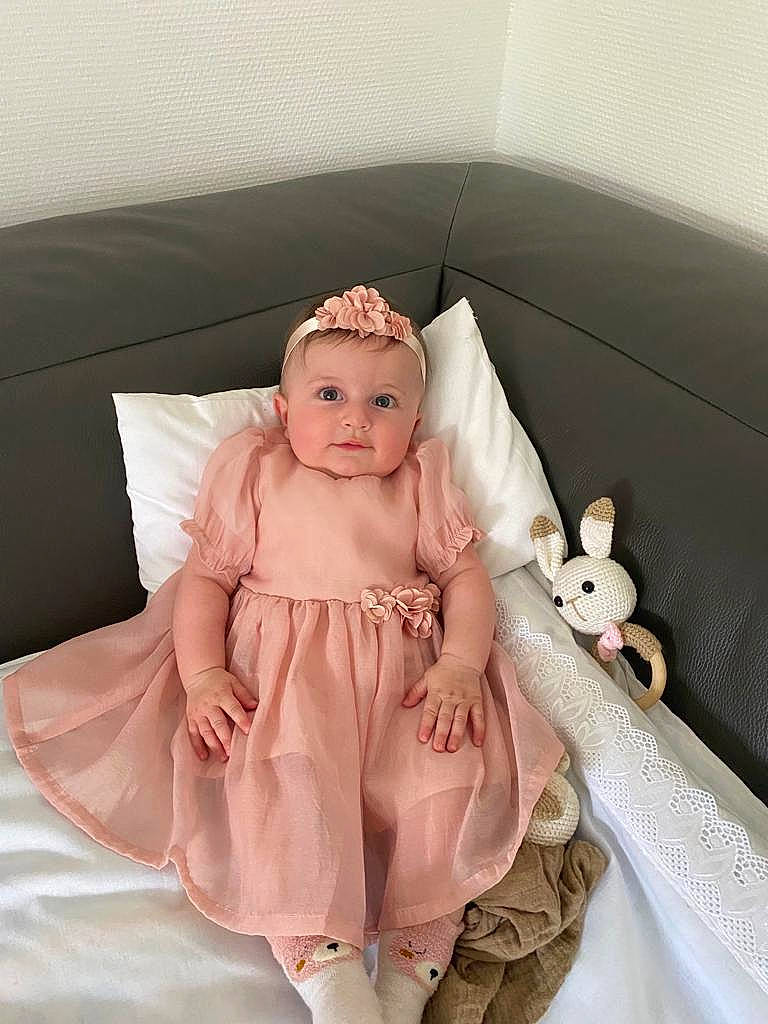 Ela participe au concours pour gagner de l'argent avec cette photo : baby, baby_products, baby_toddler_clothing, comfort, doll, dress, embellishment, headgear, headpiece, headwear, jewellery, linens, outerwear, pattern, peach, person, pink, ruffle, sitting, sleeve
