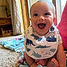 baby, infant, smiling, happy, bib, dinosaur_print, blanket, carpet, indoors, natural_light, window, curtain, furniture, lamp, wooden_furniture, feet, cute, child, portrait, home