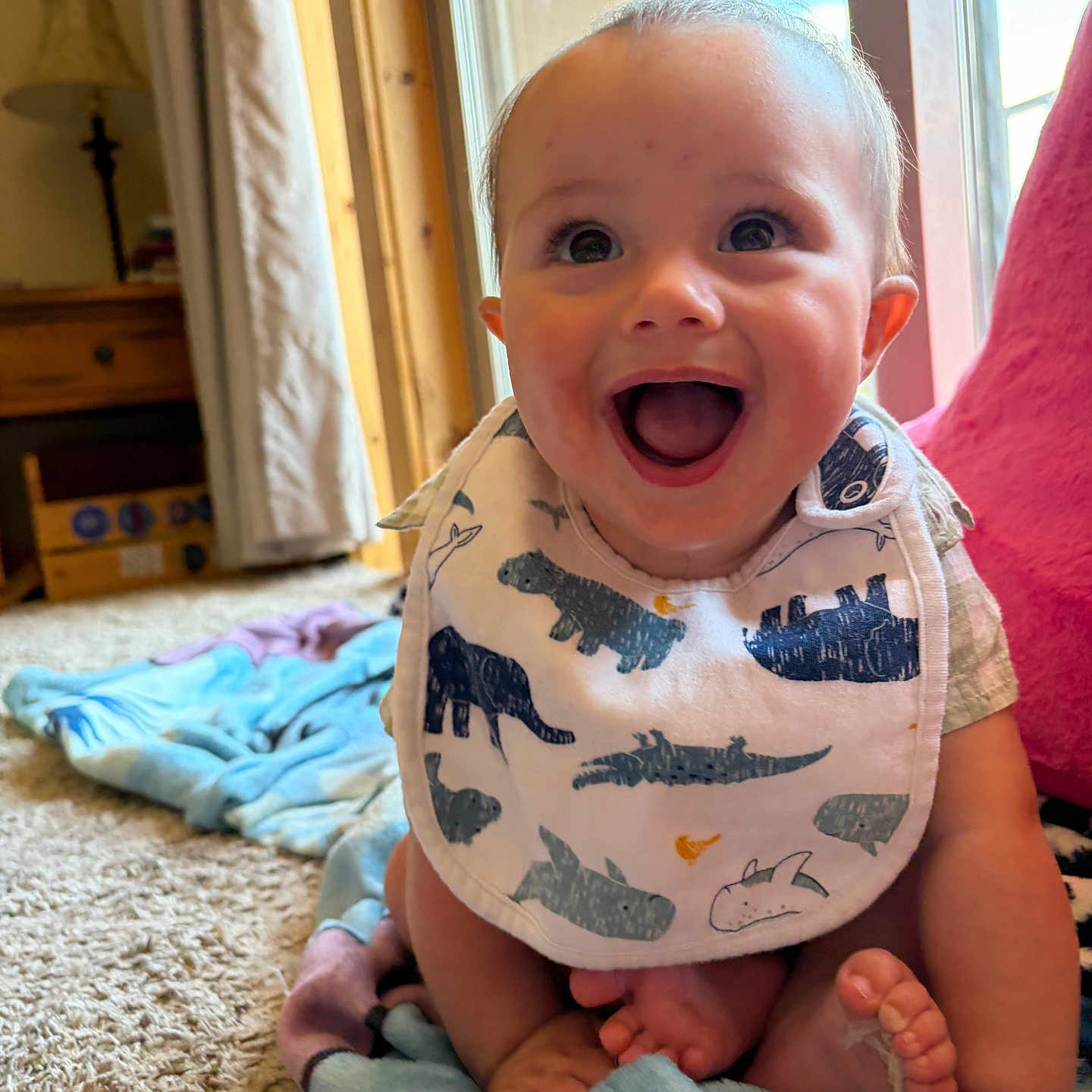 Rylan joined the competition — help win amazing prizes! baby, infant, smiling, happy, bib, dinosaur_print, blanket, carpet, indoors, natural_light, window, curtain, furniture, lamp, wooden_furniture, feet, cute, child, portrait, home
