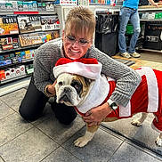 Moose is registered to the contest to win money with this photo: woman, dog, bulldog, christmas_costume, santa_hat, indoor, store, tiled_floor, shelves, tobacco_products, smiling, glasses, casual_clothing, blue_jeans, sweater, watch, kneeling, pet, holiday, festive