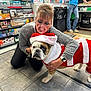 woman, dog, bulldog, christmas_costume, santa_hat, indoor, store, tiled_floor, shelves, tobacco_products, smiling, glasses, casual_clothing, blue_jeans, sweater, watch, kneeling, pet, holiday, festive