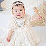 baby, child, dress, headband, toy, wooden_toy, smile, happy, indoor, fairy_lights, soft_lighting, portrait, cute, fluffy_rug, white, decor, infant, sitting, curious, adorable