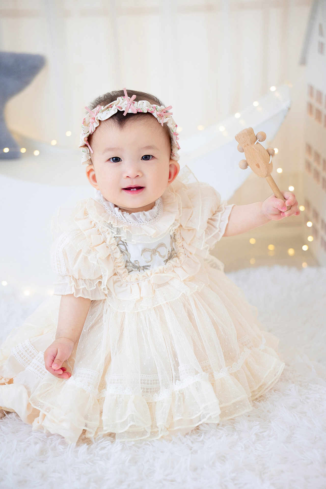 Sara is registered to the contest to win money with this photo: baby, child, dress, headband, toy, wooden_toy, smile, happy, indoor, fairy_lights, soft_lighting, portrait, cute, fluffy_rug, white, decor, infant, sitting, curious, adorable